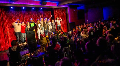 Performers standing on a brightly lit stage with red curtains and blue lighting, facing a lively audience capturing the moment with phones in a casual entertainment venue.