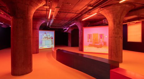 Industrial-style interior with concrete columns, exposed ceiling pipes, warm orange-red lighting, and multimedia projections showcasing artistic installations in a modern exhibition space.  Image Credit: Alex Da Corte, Mouse Museum (Van Gogh Ear), 2024. Ear Worm, installation view, MOCA Toronto, 2024. Photograph: LF Documentation.