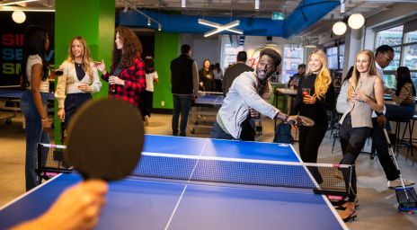 People playing table tennis in a lively indoor recreational space, surrounded by spectators holding drinks, with green and blue walls and bright overhead lighting