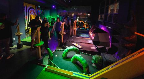 Indoor mini golf course with colorful snake-like obstacles, dim lighting, and people playing or watching in a vibrant, playful atmosphere.