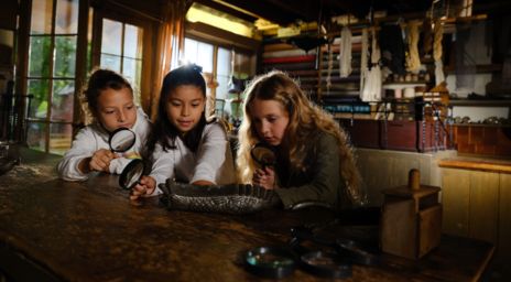 Three children closely examining a metallic artifact on a rustic wooden table using magnifying glasses, in a dimly lit room with shelves and natural light from a window, suggesting a historical or educational setting.