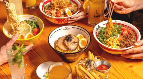 A table filled with four bowls of ramen featuring various toppings like boiled eggs, corn, pork, and vegetables, accompanied by drinks including Sapporo beer, a mint cocktail, and orange juice, with a side of fries and visible hands using chopsticks.