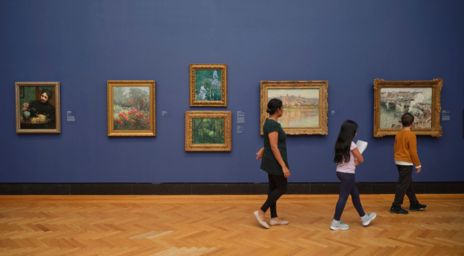Art gallery with framed paintings on a blue wall and visitors walking by.