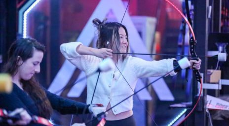 Two people practicing archery indoors with modern lighting.