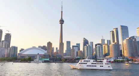 The Toronto skyline with the CN Tower and Rogers Centre, viewed from a waterfront with boats, including a large white yacht, on rippled water under a clear blue sky.