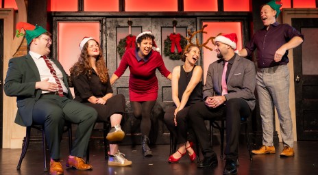 Festive group of six people wearing holiday accessories, laughing together on stage.