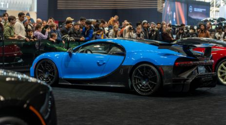 A blue exotic car on display at the AutoShow with several attendees surrounding space and taking photos.